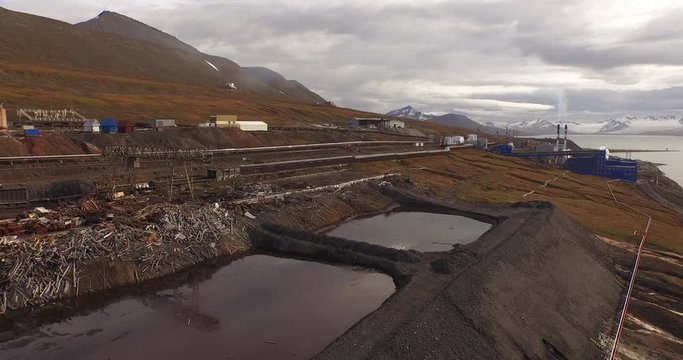 Barentsburg. The aerial panoramic view over the industrial part of the northern mining settlement in Svalbard. The harsh nature of the arctic coastline. Cloudy sky and snowy mountains in a distance.4K