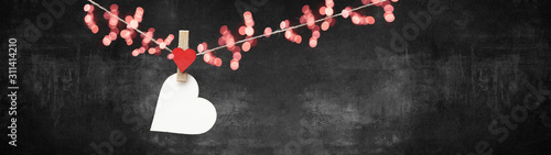 Happy Valentine's Day background banner panorama - White heart hang on wooden clothes pegs with wooden heart and bokeh lights on a string isolated on black concrete stone wall, with space for text