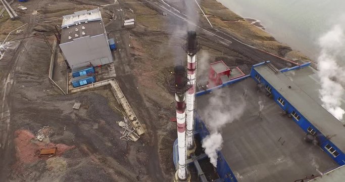 The aerial view over the two long smoking factory pipes of industrial building standing at the coast of the Arctic ocean. Technical constructions scattered around on the shore. Barentsburg.Svalbard.4K