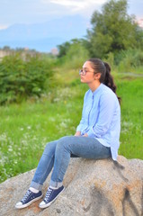 Naklejka na ściany i meble A young girl wearing glasses in a blue shirt and jeans sits on a large stone in a green clearing. Selective focus.