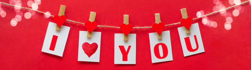 I love you - Clothes pegs with wooden hearts, bokeh lights and paper notes hang on rope isolated on red texture background banner panorama long