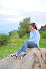 Fototapeta na wymiar A young girl wearing glasses in a blue shirt and jeans sits on a large stone in a green clearing. Selective focus.