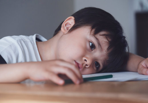 Kid playing colour pencil sitting alone and looking down with bored face, Preschool child boy laying head down on table looking out deep in thought, Five year old kid bored with school homework