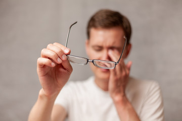 the young man holds glasses with diopter lenses and rubs his eyes from fatigue, fatigue of vision and eyes, computer vision syndrome, the problem of myopia, vision correction