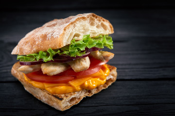 Appetizing sandwich on a wooden board. Baguette sandwich with filling from lettuce, slices tomato. Dark wooden background. View from above. Close-up. Macro photography.