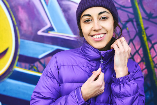 Close-up portrait of young smiling woman wearing in purple down jacket and knitted hat posing against wall painted with graffiti. Urban clothing style, urban background.