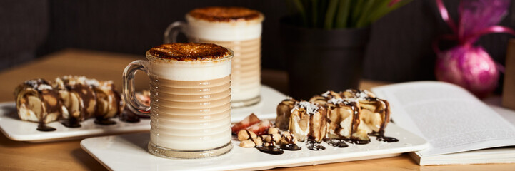 Banner size image of two cups of Hot latte with baked caramel crust and sweet roll with banana and strawberry on the wooden table in coffe-shop. Coffee concept