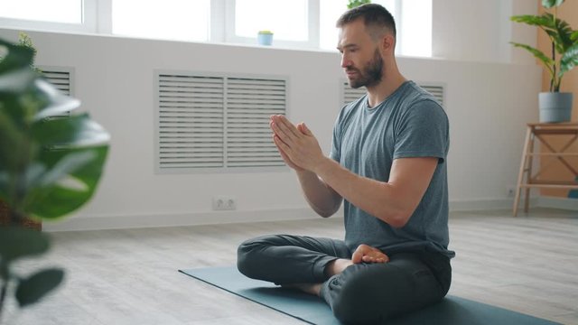 Slow motion of serious young man meditating at home moving hands in namaste with closed eyes sitting on yoga man in cozy apartment. People and relaxation concept.