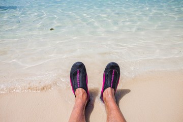 Close up view of rolling waves on men's feet standing on white sand beach in swimming shoes .