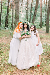 Obraz na płótnie Canvas Pretty bride and two bridesmaids in white dresses smiling and looking at camera while standing on the background of pine forest. Stylish and rustic wedding. Marriage concept