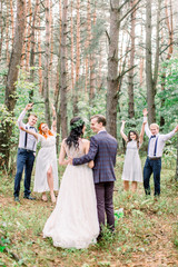 Fototapeta premium Stylish rustic wedding couple with bridesmaids and groomsmen at forest on wedding day. Back view of groom and bride, embracing and kissing. Their friends smiling and holding their hands on the air.