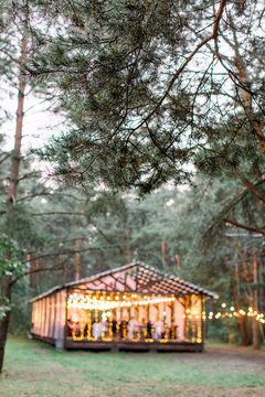 Blurred photo of wooden tent restaurant with wooden tables and chairs., decorated with light bulbs Vintage design of the restaurant in the forest in summer. Wedding reception in the open air.