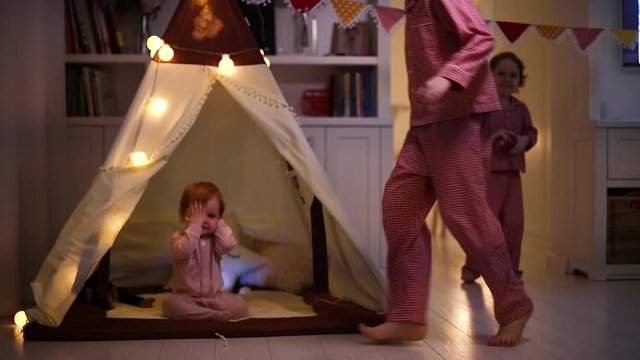 laughing kids, siblings having fun, running and playing around a teepee tent in the evening at home
