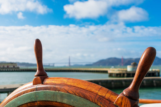 Close up. Ship steering wheel with blurred pier, Golden Gate Bridge and seascape background on the cloudy horizon