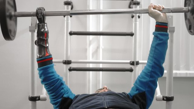 An athlete without a hand raises the barbell while lying down. Holds the bar with a professional sports hand prosthesis. Gym for Paralympics