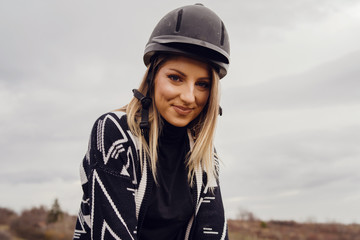 Young beautiful blonde caucasian woman female portrait against a gray cloudy sky in winter or autumn day wearing protective black helmet and sweater