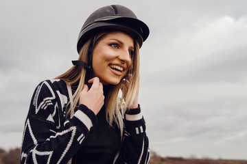 Young beautiful blonde caucasian woman female adjusting black protective helmet against a cloudy gray sky in winter or autumn day wearing sweater smiling