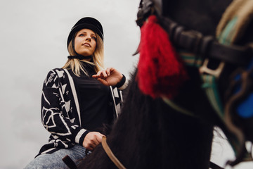 Young beautiful blonde caucasian woman female portrait against a gray cloudy sky in winter or autumn day wearing protective black helmet and sweater riding the horse low angle view