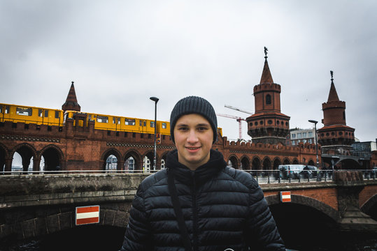 Young man smilling with Oberbaum Bridge and yellow train on background in cloudy weather in Berlin. Travel and tourism in Berlin concept. Europe travel in winter. Student and youth travel concept.