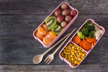 Lunch box with fresh fruits and vegetables on a dark wood background. Kiwi, tangerines, grapes, broccoli, corn, cabbage, carrots. Healthy food at work concept