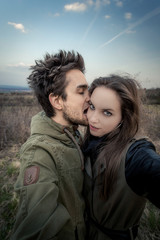 Portrait of young man, who is biting woman's face. Woman is looking in to camera. Couple is stretching hands out of picture. Background out of focus with withered vegetation and blue sky.