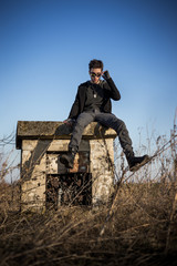 Disheveled charismatic young man in black colored outfit, with roguish expression, sitting on abandoned concrete block of installation with pure blue sky in background and withered vegetation around.