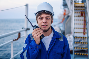 Marine Deck Officer or Chief mate on deck of offshore vessel or ship , wearing PPE personal protective equipment - helmet, coverall. He holds VHF walkie-talkie radio in hands.