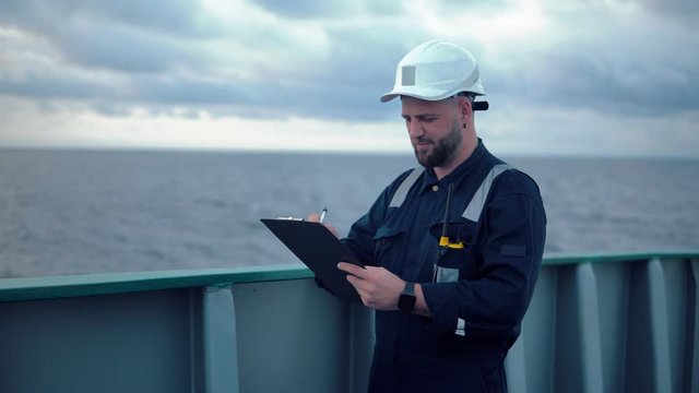 Marine chief officer or chief mate on deck of ship or vessel. He fills up ahts vessel checklist. Ship routine paperwork. He holds VHF walkie-talkie radio in hands.