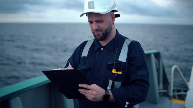 Marine chief officer or chief mate on deck of ship or vessel. He fills up ahts vessel checklist. Ship routine paperwork. He holds VHF walkie-talkie radio in hands.