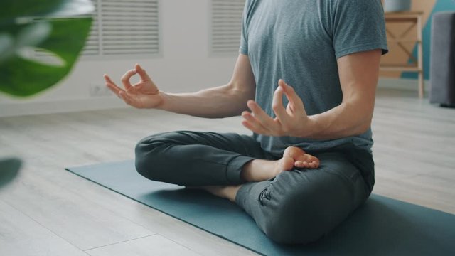 Low shot of anonymous man meditating on yoga mat sitting in lotus position with hands in namaste then in mudra gestures. People and relaxation concept.