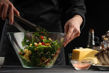 Cooking salad, mixing in a large transparent plate. On a black background with ingredients. Cooking, gastronomy, home recipes. Recipe book, delicious food concept. Home cooking, Greek or Italian salad
