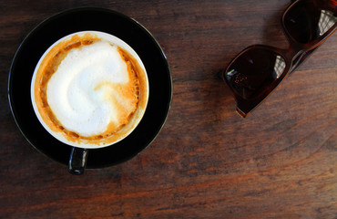 Top view or flat lay of hot cappuccino in black cup with brown sunglasses and on brown wooden background or table with copy space. Refreshment drinking with coffee at cafe shop in the morning time.
