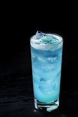 electric light blue drink with ice and blue garnish served in a tall clear glass with all black background 