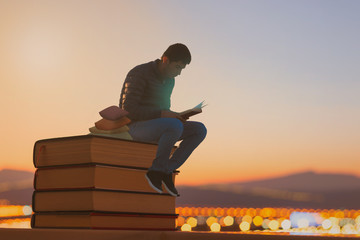 a young boy reading a book on stack of books outdoors with the view of mountains at sunset.