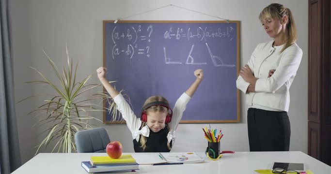 Little Caucasian girl sitting at the table in headphones and showing thumbs up as adult tutor standing next to her. Schoolgirl misbehaving studying at home. Education, learning. Cinema 4k ProRes HQ.