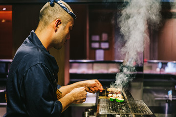 Japanese Yakitori Chef is grilling chicken marinated with ginger, garlic and soy sauce and cucumber with a lot of smoke.