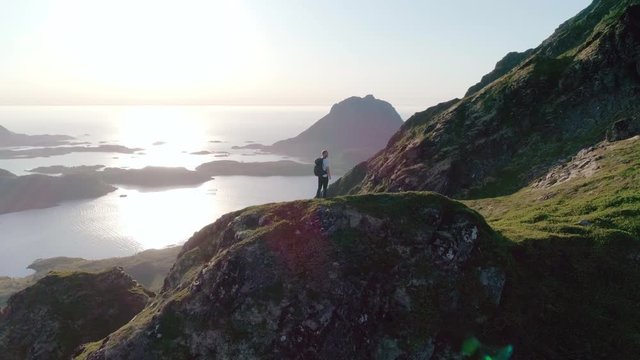Wide 4K slow motion drone shot with orbit motion of Norwegian climber hiker in 30s with backpack standing on mountain side admiring the stunning panoramic sunset view in Lofoten Vester&aring;len in Norway.