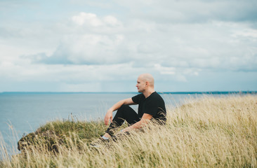 A bald man in black shirt sitting on grass on the edge of the rocky coast of the Baltic sea.