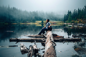 An Asian woman sitting down and looking up reflecting and pondering in the middle of foggy mountain at a lake - obrazy, fototapety, plakaty