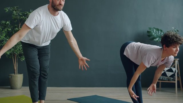 Tilt-up slow motion of man and woman young couple doing yoga in cozy studio bending body then raising arms standing on mats. Youth, healthy lifestyle and people concept.