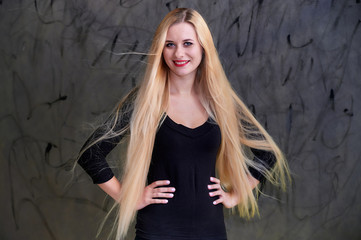 Concept of a young blonde woman with a chic hairstyle. Portrait of a cute girl in a black T-shirt with long beautiful hair and great makeup. Smiling, showing emotions on a gray background.