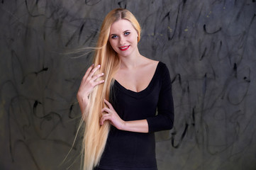 Concept of a young blonde woman with a chic hairstyle. Portrait of a cute girl in a black T-shirt with long beautiful hair and great makeup. Smiling, showing emotions on a gray background.