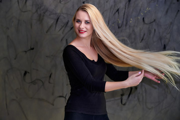 Obraz premium Concept of a young blonde woman with a chic hairstyle. Portrait of a cute girl in a black T-shirt with long beautiful hair and great makeup. Smiling, showing emotions on a gray background.