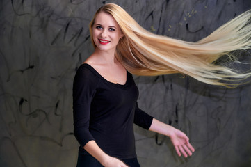 Naklejka premium Concept of a young blonde woman with a chic hairstyle. Portrait of a cute girl in a black T-shirt with long beautiful hair and great makeup. Smiling, showing emotions on a gray background.