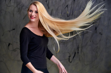 Concept of a young blonde woman with a chic hairstyle. Portrait of a cute girl in a black T-shirt with long beautiful hair and great makeup. Smiling, showing emotions on a gray background.