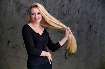 Concept of a young blonde woman with a chic hairstyle. Portrait of a cute girl in a black T-shirt with long beautiful hair and great makeup. Smiling, showing emotions on a gray background.