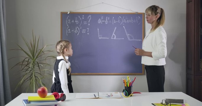 Portrait of smart Caucasian woman explaining math to cute little girl. Tutor pointing at geometrical formulas written on blackboard and talking. Teaching, education concept. Cinema 4k Prores HQ.