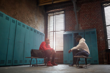 Two African men in sports clothing sitting on bench and talking to each other in locker room
