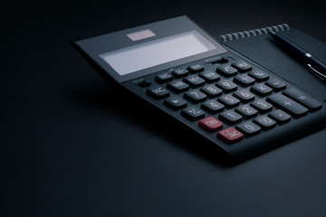 Side view of a black accounting calculator with blank screen, pen and notebook on a dark wooden office desk with copy space. Business and finance template.  Tax and profit calculation concept - obrazy, fototapety, plakaty