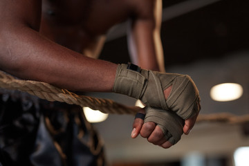 Fototapeta premium Close-up of African boxer in bandage on his hands leaning on the ropes of boxing ring in the gym